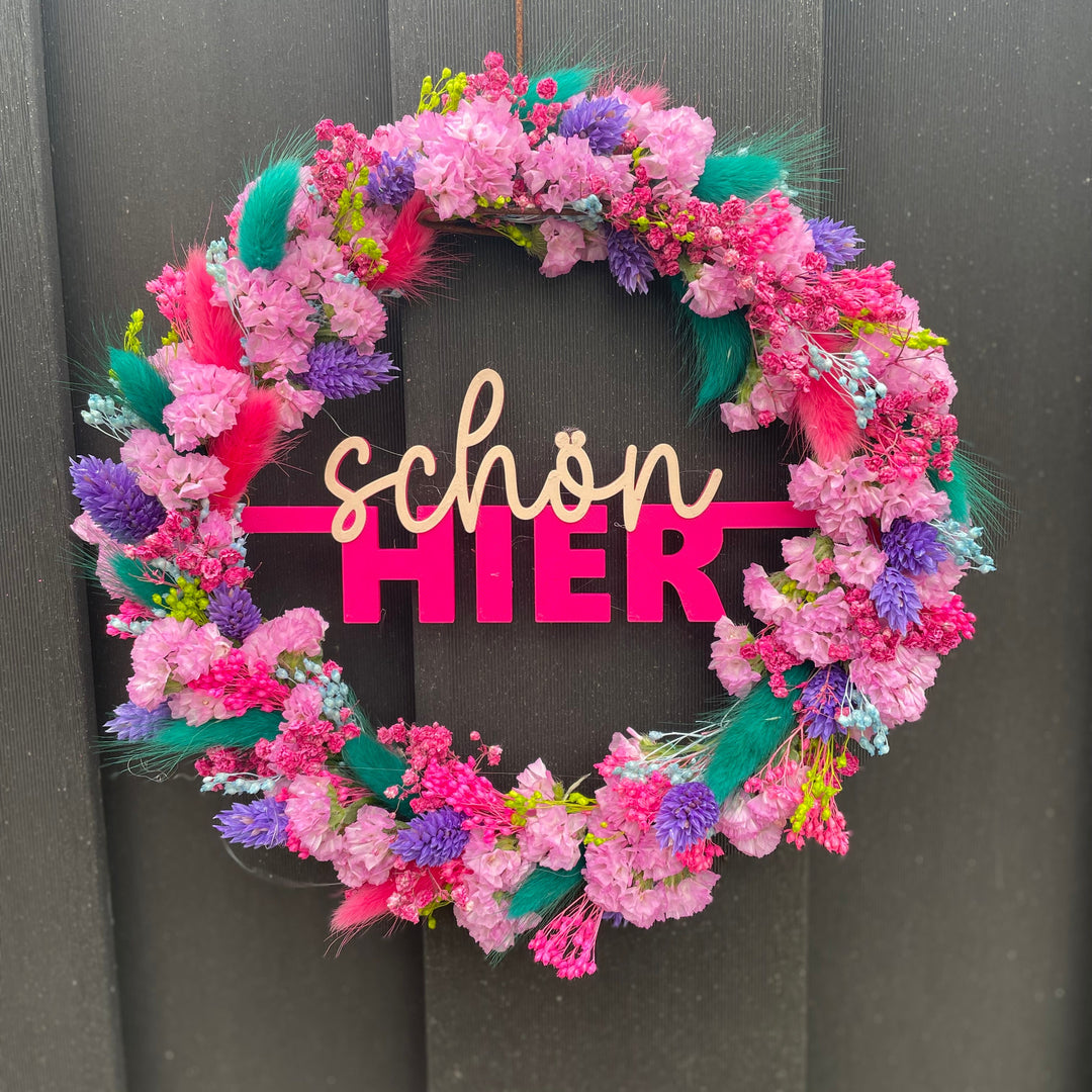 Pinker Kranz mit "schön hier" als Schriftzug in der Mitte umwickelt mit bunten Trockenblumen