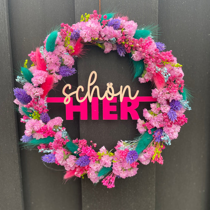 Pinker Kranz mit "schön hier" als Schriftzug in der Mitte umwickelt mit bunten Trockenblumen